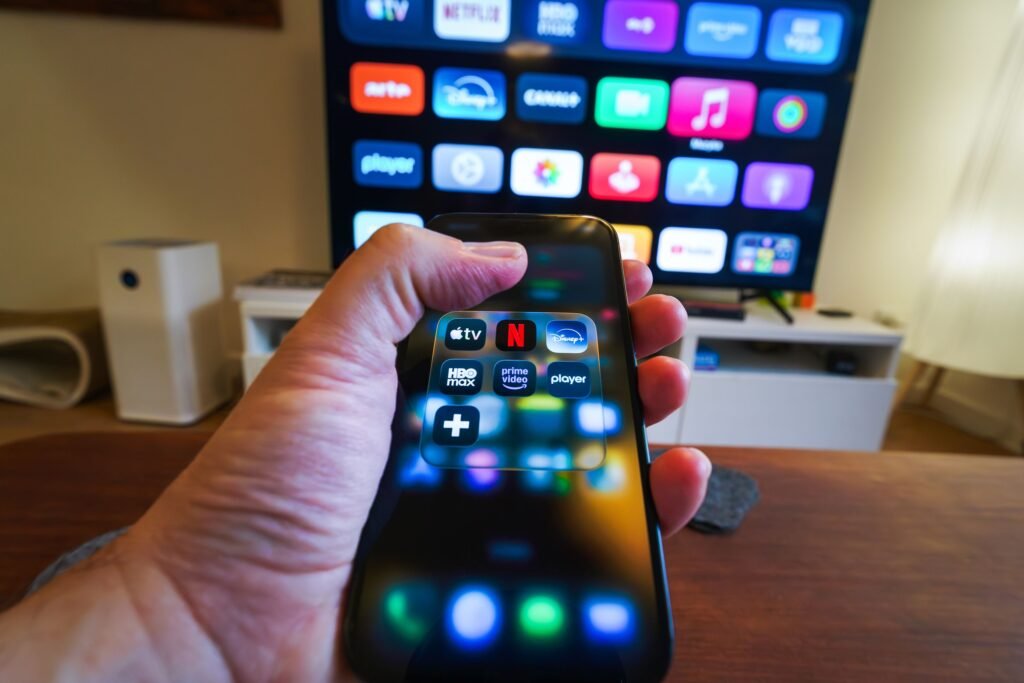 $219 a Month on Subscriptions: Do This 10-Minute Audit Before Your July Bills Hit Close-up of a hand holding a phone displaying streaming apps in front of a TV with multiple app icons.